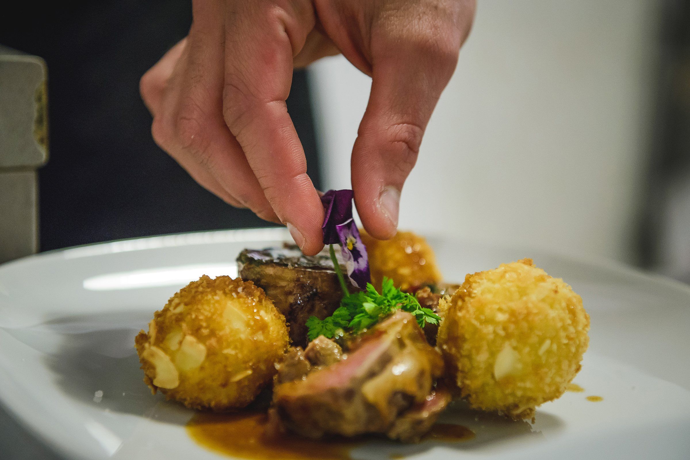A hand placing a purple edible pansy on a plated dish, golden almond-crusted croquettes, glazed meat in a jus and a sprig of parsley on a white plate.