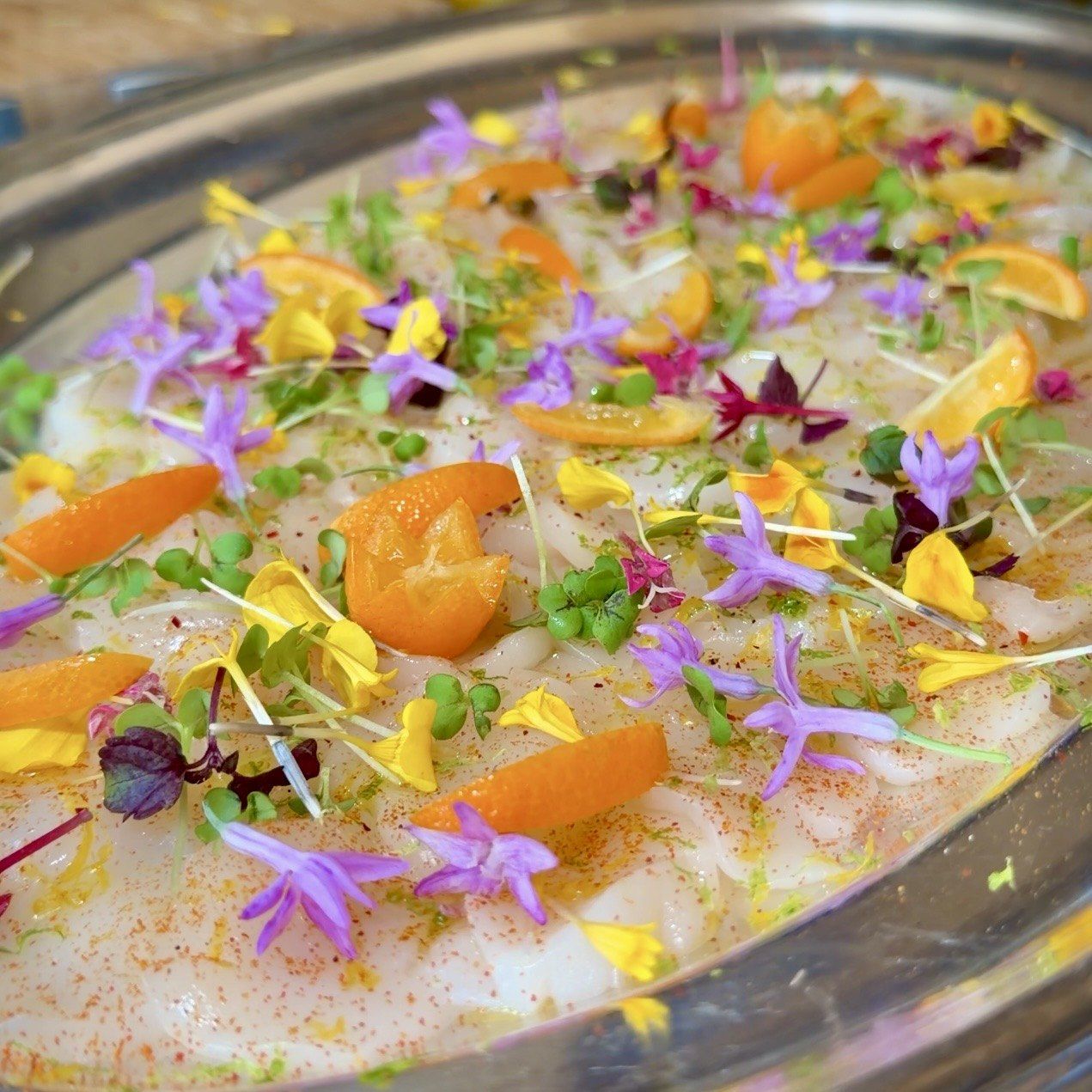 Carpaccio en fines tranches, quartiers d’agrumes, jeunes pousses et fleurs comestibles violettes et jaunes.
