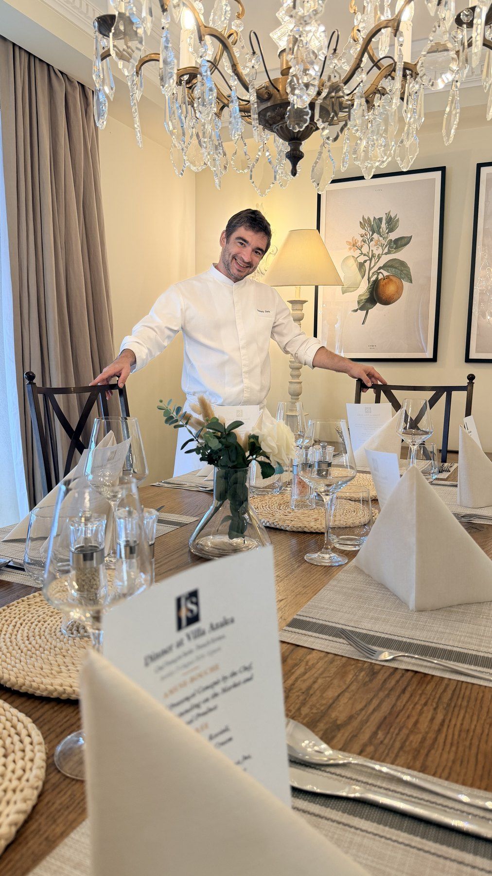 Le chef François Savin devant une table dressée sous un lustre en cristal, menus imprimés à chaque couvert.