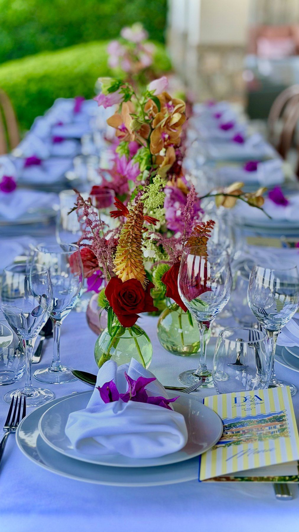 Longue table dressée en extérieur, grand bouquet de fleurs, verrerie et serviettes pliées.