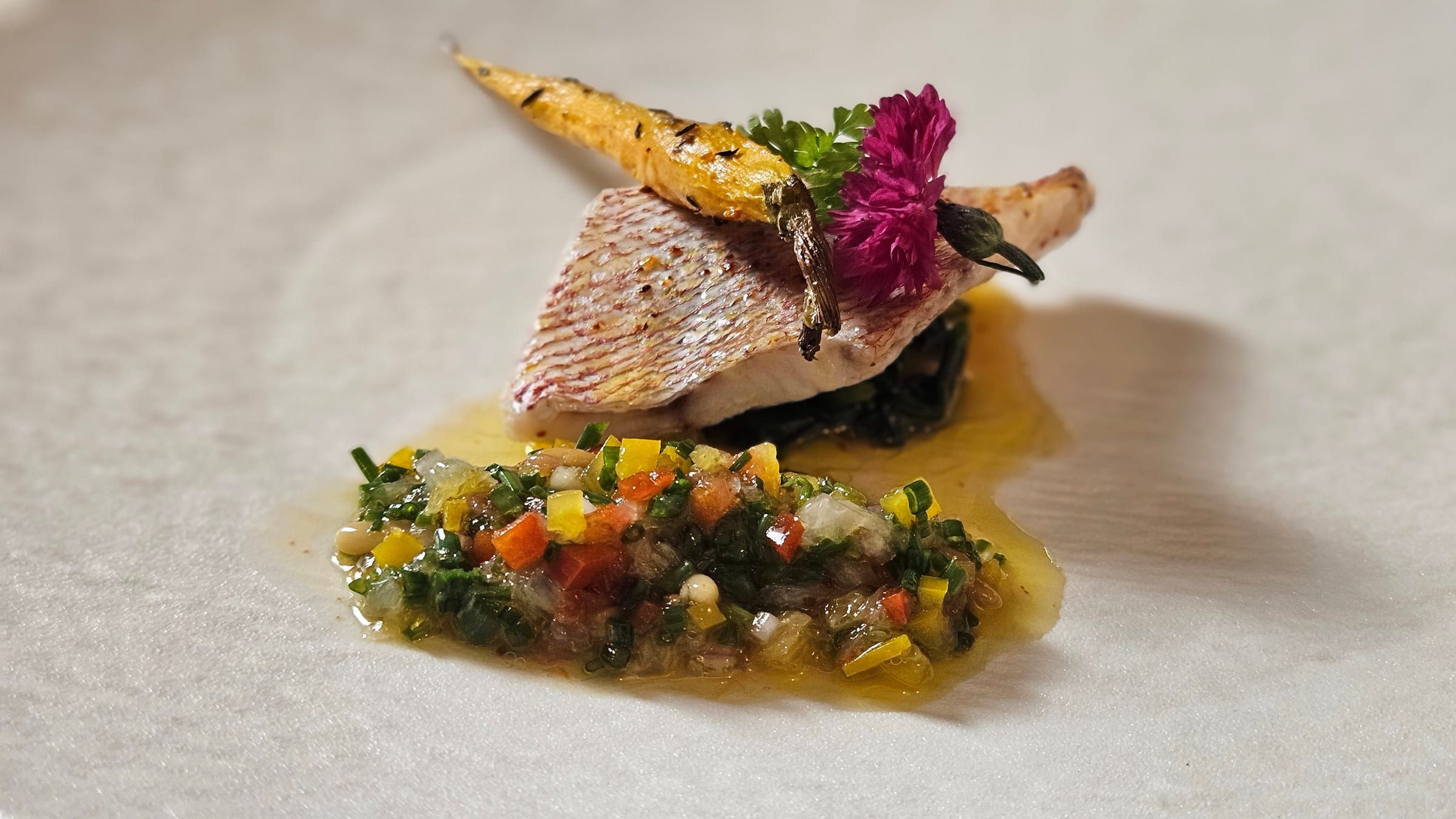 A seared fish fillet with a glazed baby carrot, a vegetable condiment and an edible flower.