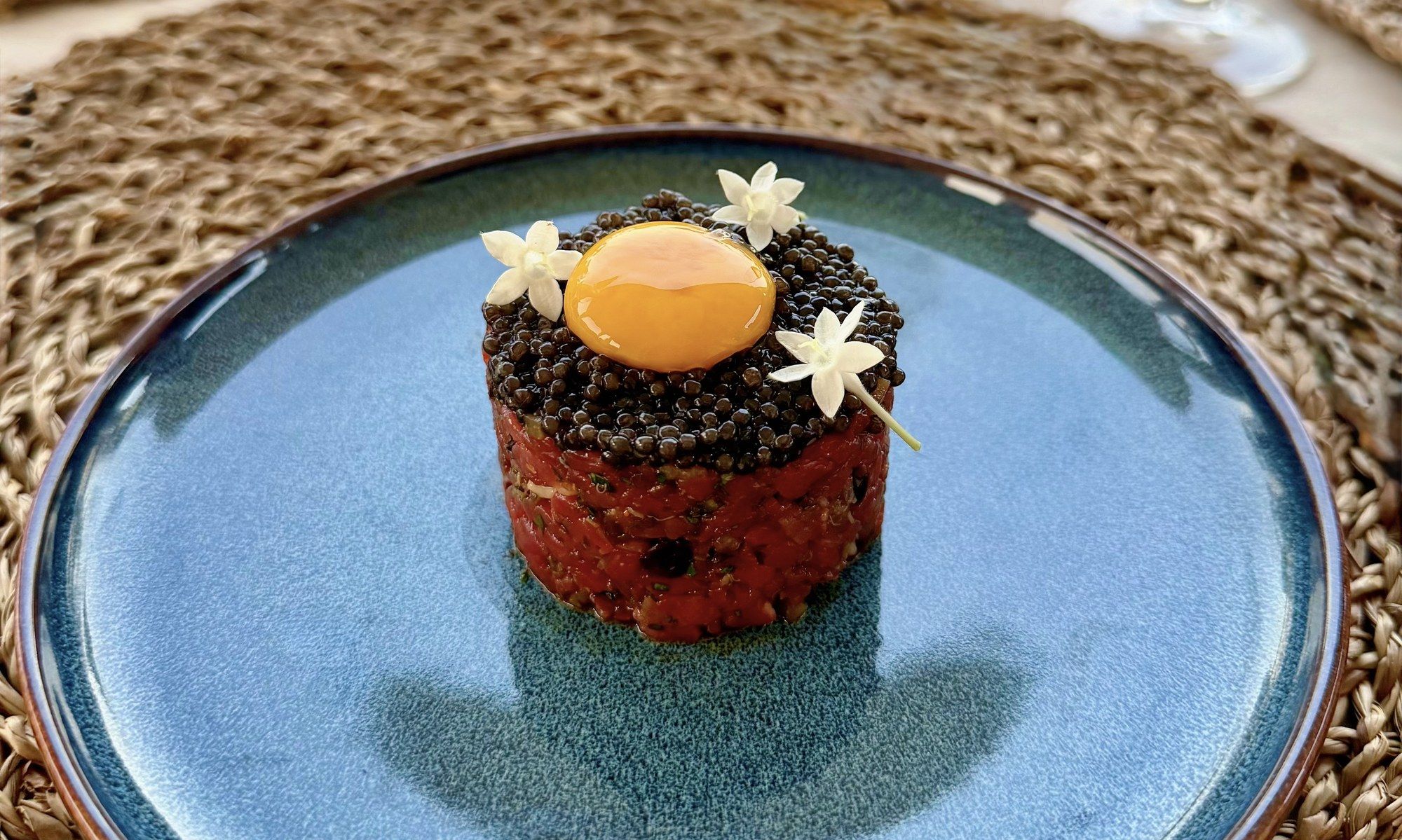 Beef tartare topped with caviar and an egg yolk, finished with white flowers, on a blue glazed plate.