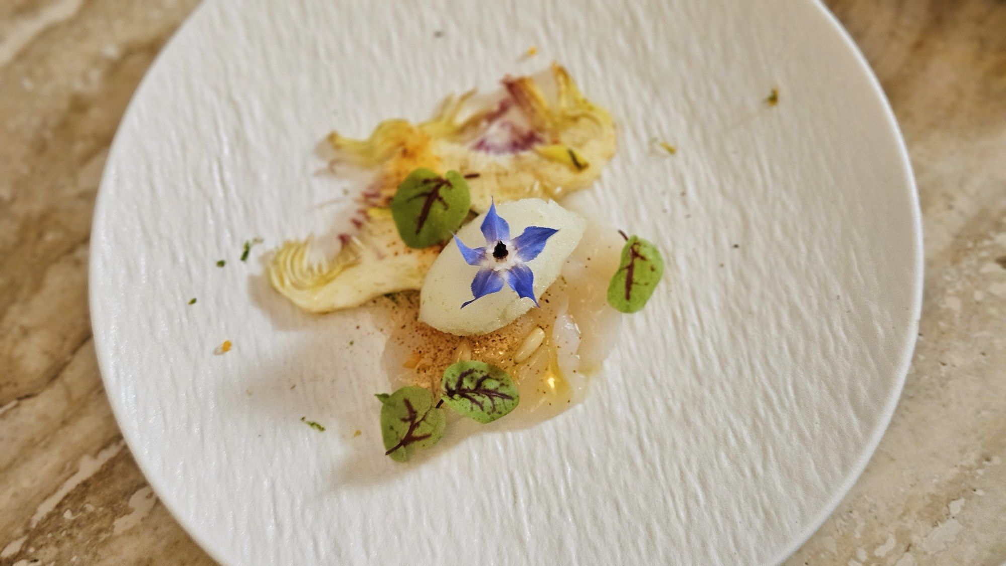 Thinly sliced carpaccio with artichoke, sorrel leaves and a borage flower on a white plate.